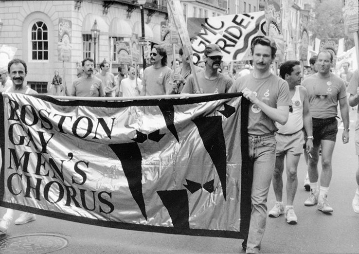 A black and white photo of a group of men walking down a street. Two men in the front hold a large banner that reads "Boston Gay Men's Chorus" while someone in the back holds a flag that says "BGMC Pride"