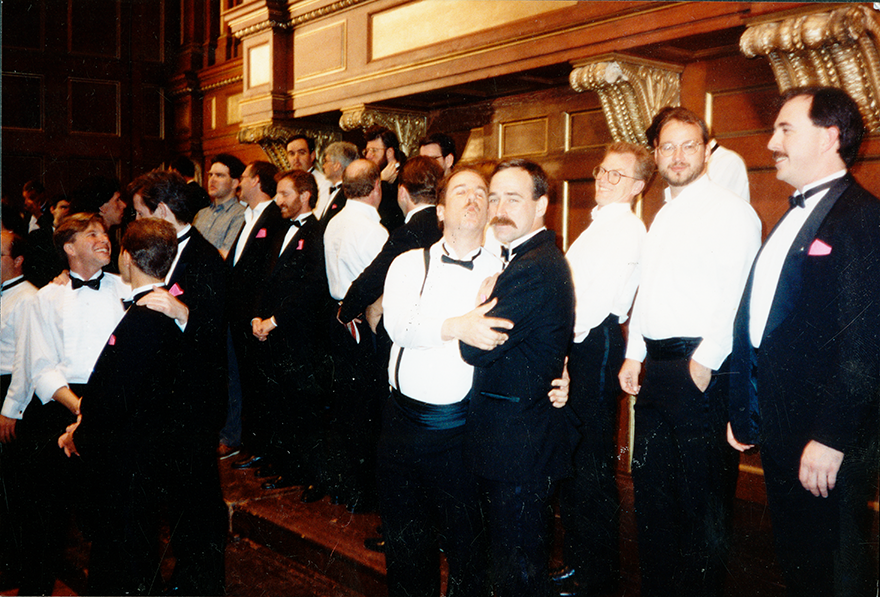 A group of men in tuxedos and dress clothes stand informally smiling and chatting. Two men in the center pose for the camera hugging and making kiss faces.