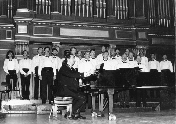 A black and white photo of a Boston Gay Men's Chorus live performance, with a view of center stage and a pianist, from 1987.