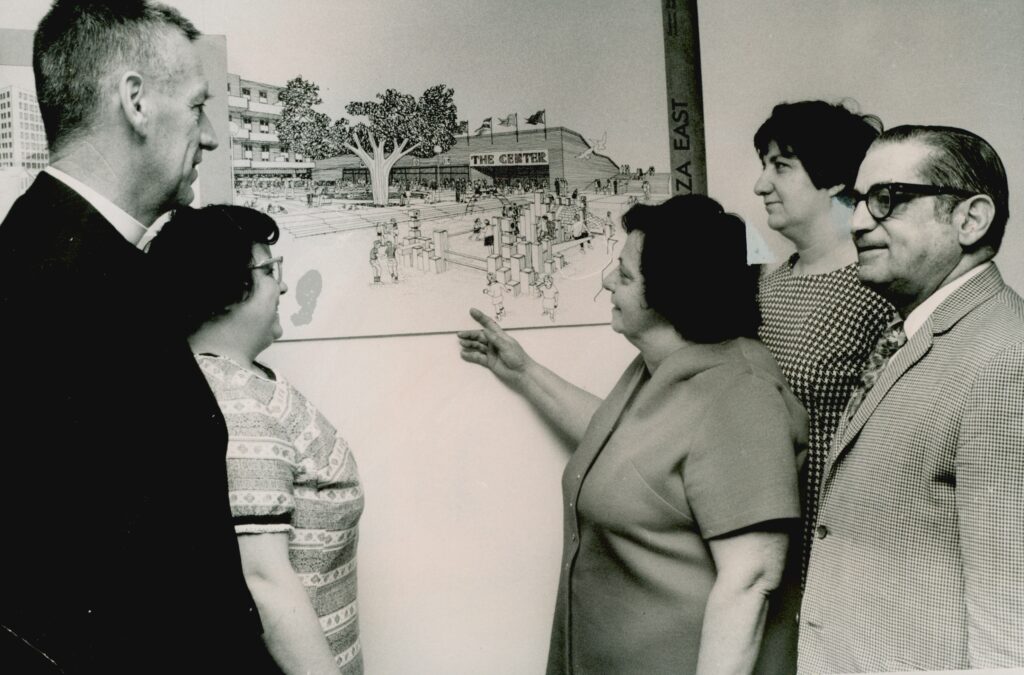 A group of people stand in front of a rendering of a community center. The woman in the center is pointing at it while the others look on