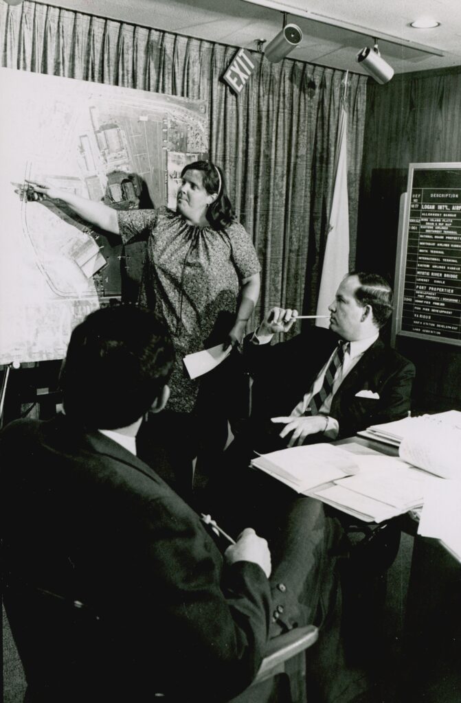 A black and white photo of a woman standing in front of city planning map and pointing while two seated men look on.