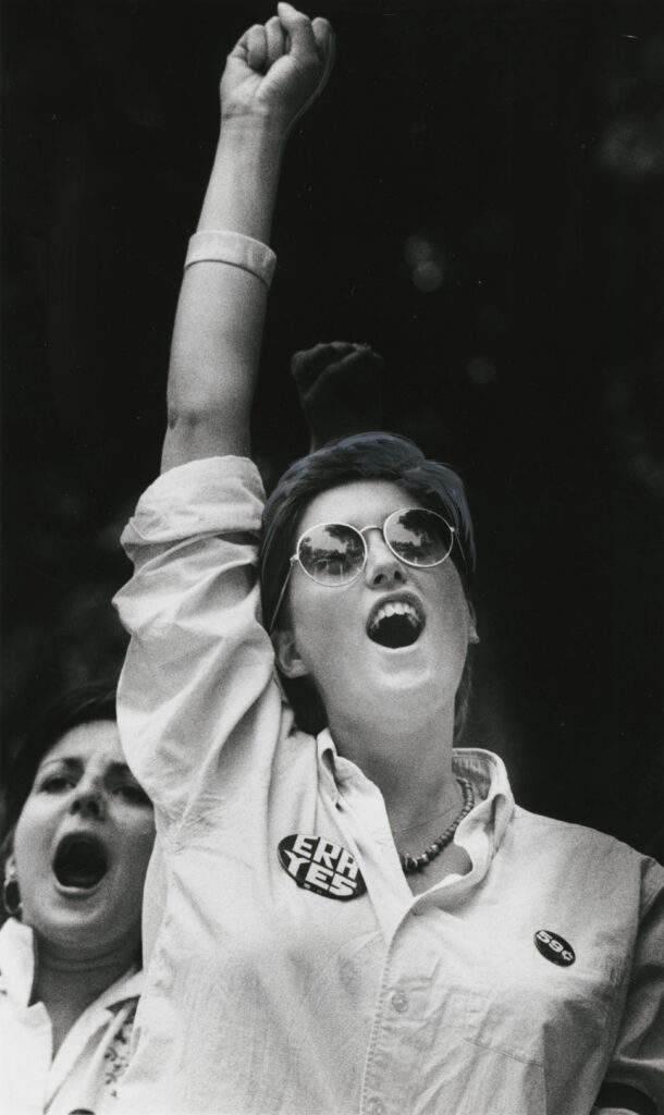 A woman wearing sunglasses, a white button-down shirt, and a large button that says "ERA YES" holds her right fist in the air and yells.