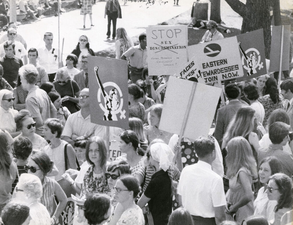 A large group of protestors hold signs with messages of women's power and urging the stoppage of sex discrimination.