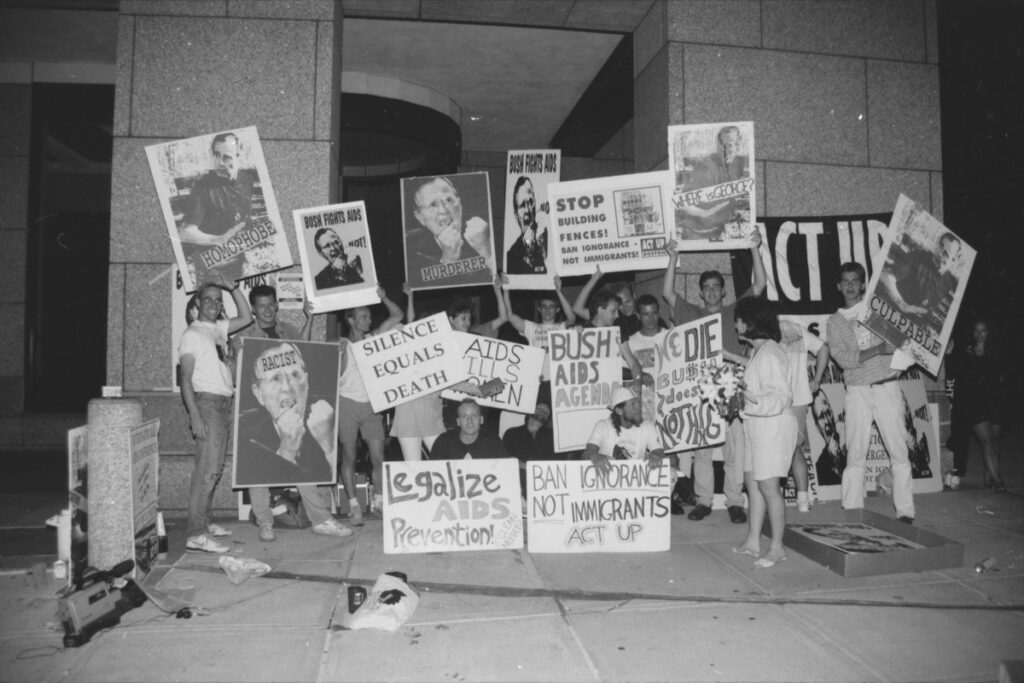 A group of protestors hold signs criticizing George HW Bush and demanding AIDS prevention and resources.