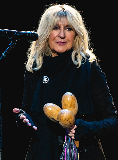 Christine McVie standing in front of a microphone holding three maracas. She has long blonde hair and is wearing black.