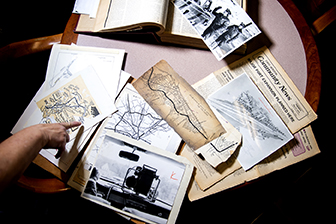Archival photos, books, documents, and papers are spread around a round pink table. A hand on the left side of the frame points to one of the documents.