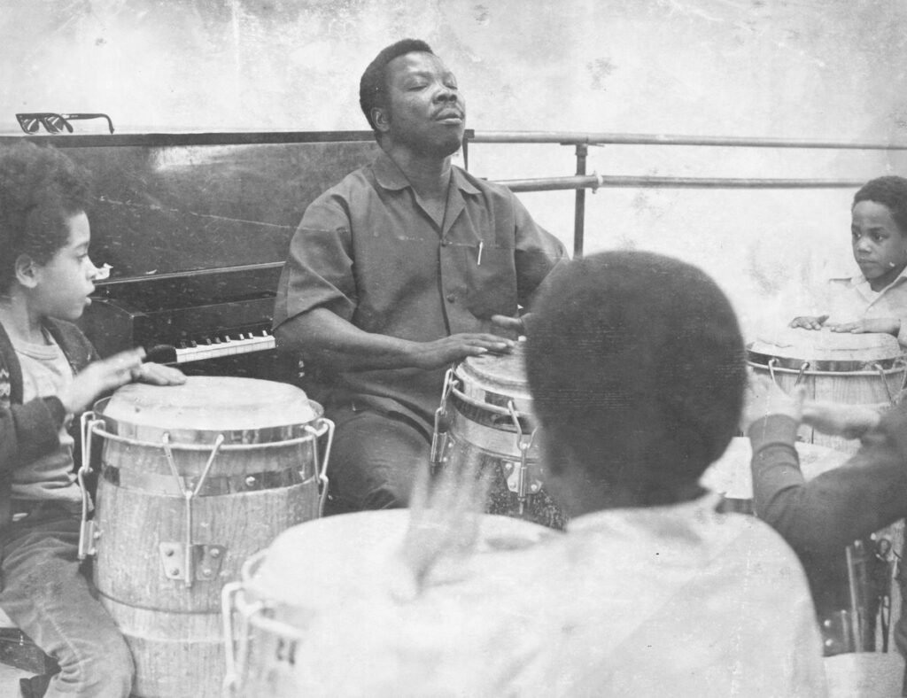 Black and white image of a Black man playing a drum with his hands. Children sit before him with their own drums.
