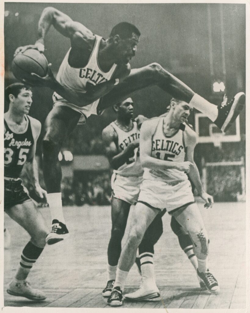 Black and white image of Bill Russell wearing a white Celtics uniform holds the ball while leaping with spread legs.