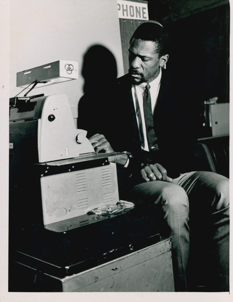 Black and white image of Bill Russell wearing a dark jacket and tie and sitting in front of a machine.