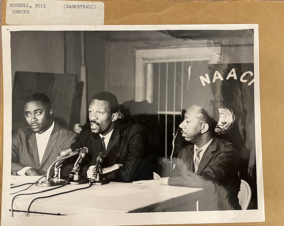 Black and white image of Bill Russell sitting at a table and speaking into several microphones. There are two other men sitting on either side of him. On a window behind him are the letters NAACP. The photo is resting on top of a folder with a label "Russell, Bill (Basketball) Groups"