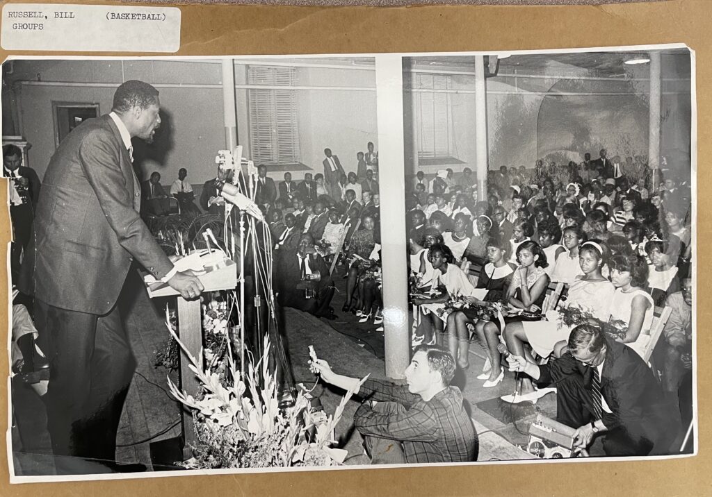 Black and white image of Bill Russell, wearing a suite and speaking to a crowd of young Black teenagers. Russell is standing on the left and facing the crowd on the right. He is so tall that he has to stoop a little to reach the microphones.