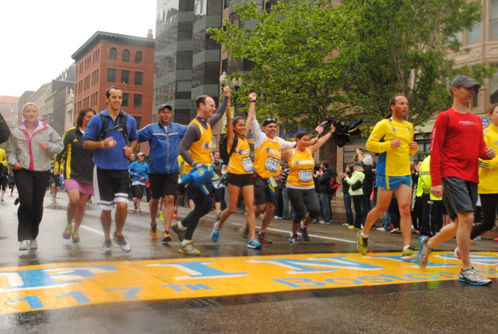 Several people run over the yellow and blue finish line of the Boston Marathon.