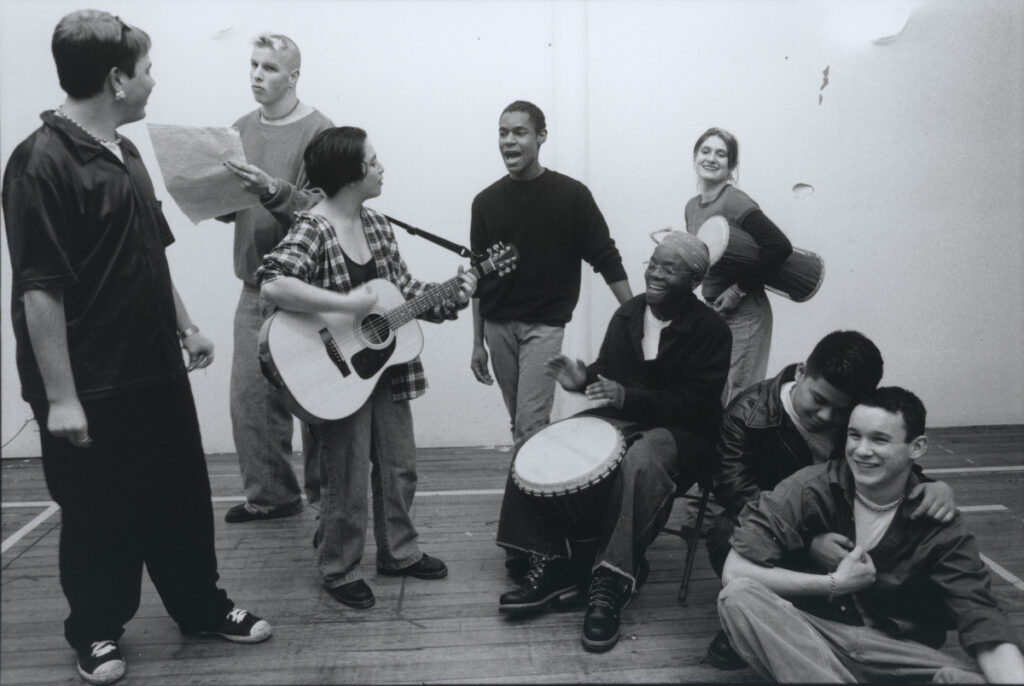 Black and white image of performers gathered together. Some are holding instruments and papers and others are singing