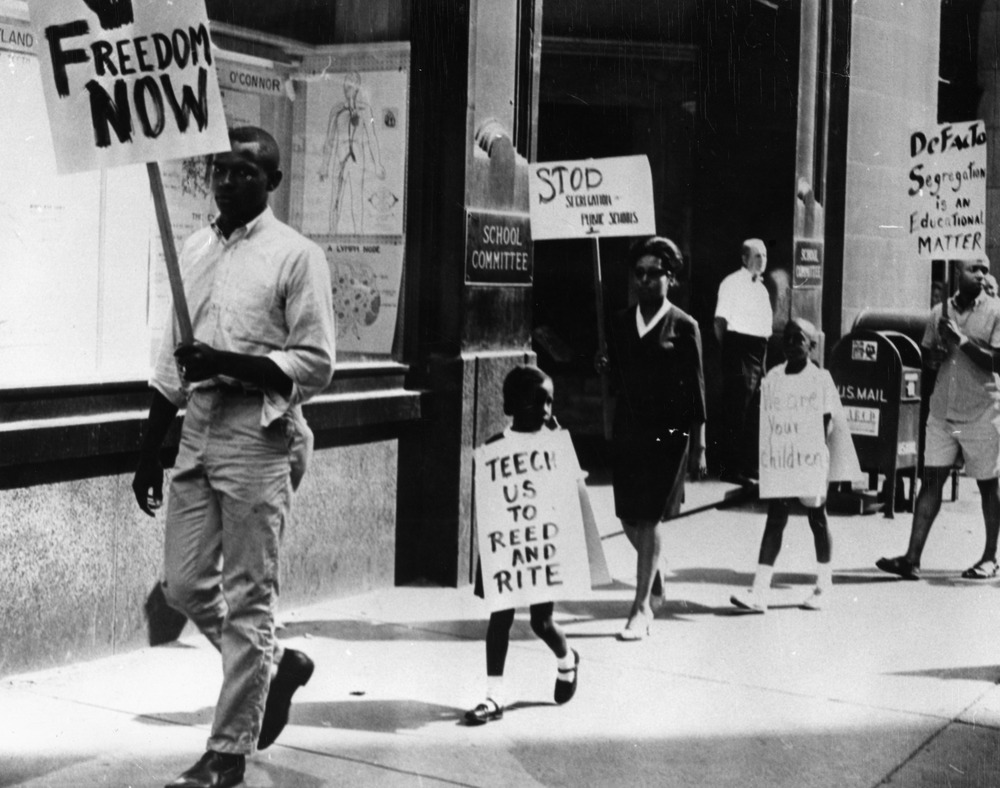 Black and white image of children and adults walking on a sidewalk holding protest signs 