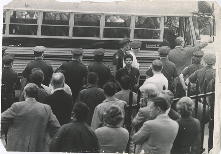Black and white image of a Black student standing in front of a school bus surrounded by police officers, with a crowd of adults looking on