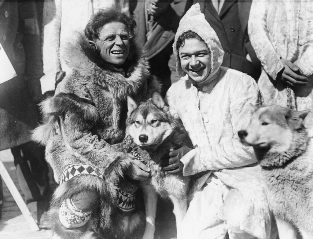 Black and white image of a husky dog. Kneeling next to him is a man wearing a fur jacket and a student in a winter coat