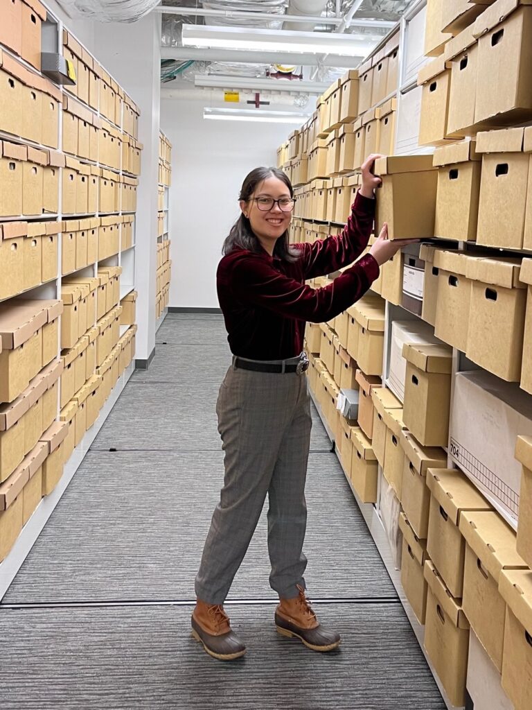 A smiling woman with glasses and a red shirt pulls a box from a shelf