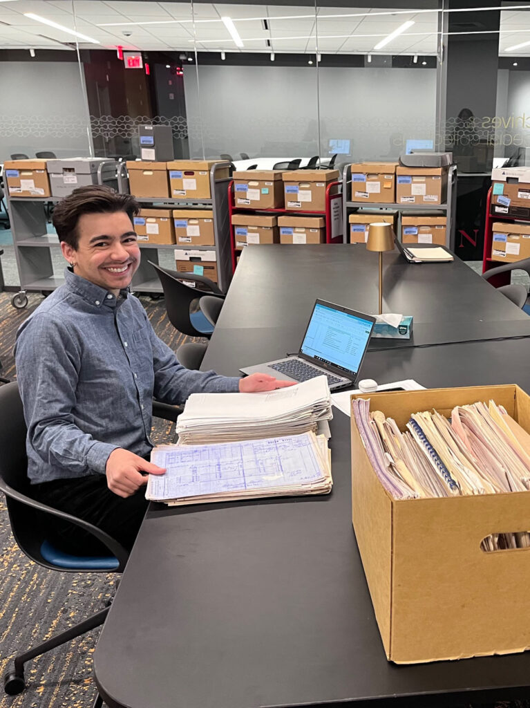 A smiling man sits at a table with a laptop and a stack of documents, surrounded by archival boxes