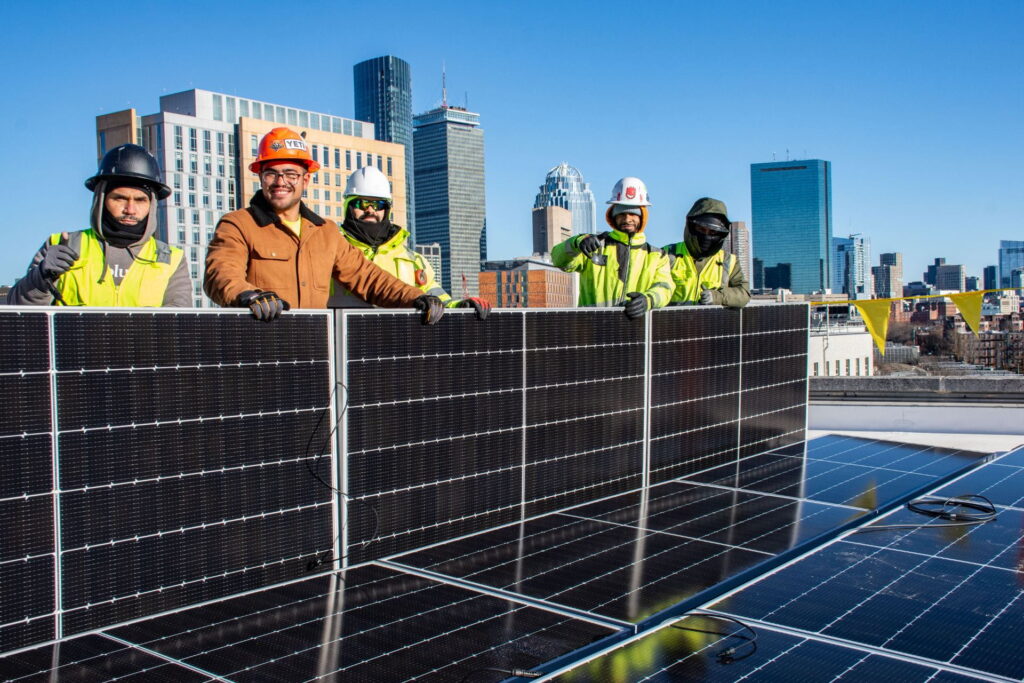 Four workers in construction gear pose with a solar panel on the roof of Snell Library