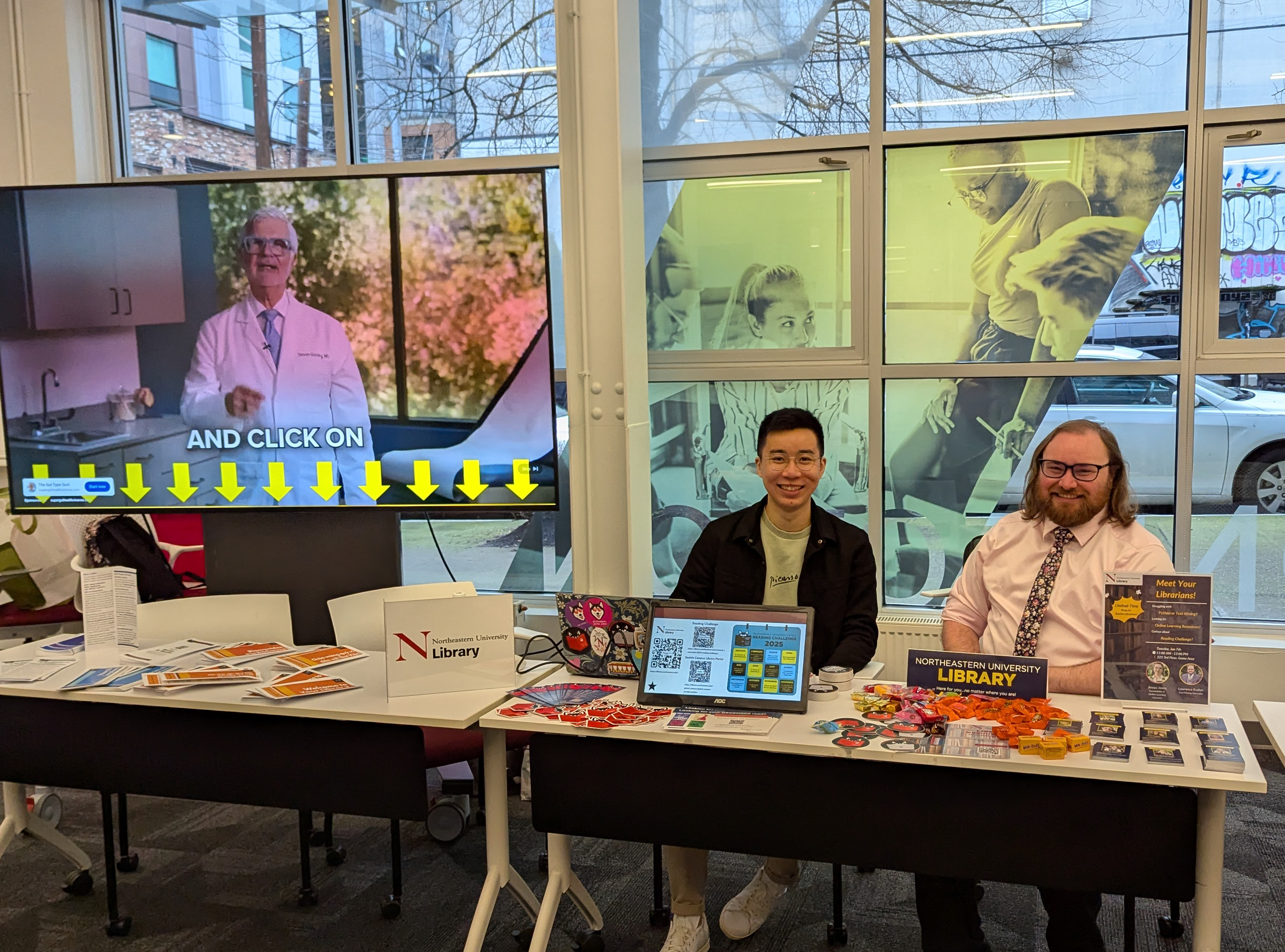 Two smiling people sit at a table covered with library handouts, candy, and computers with library content on the screens.