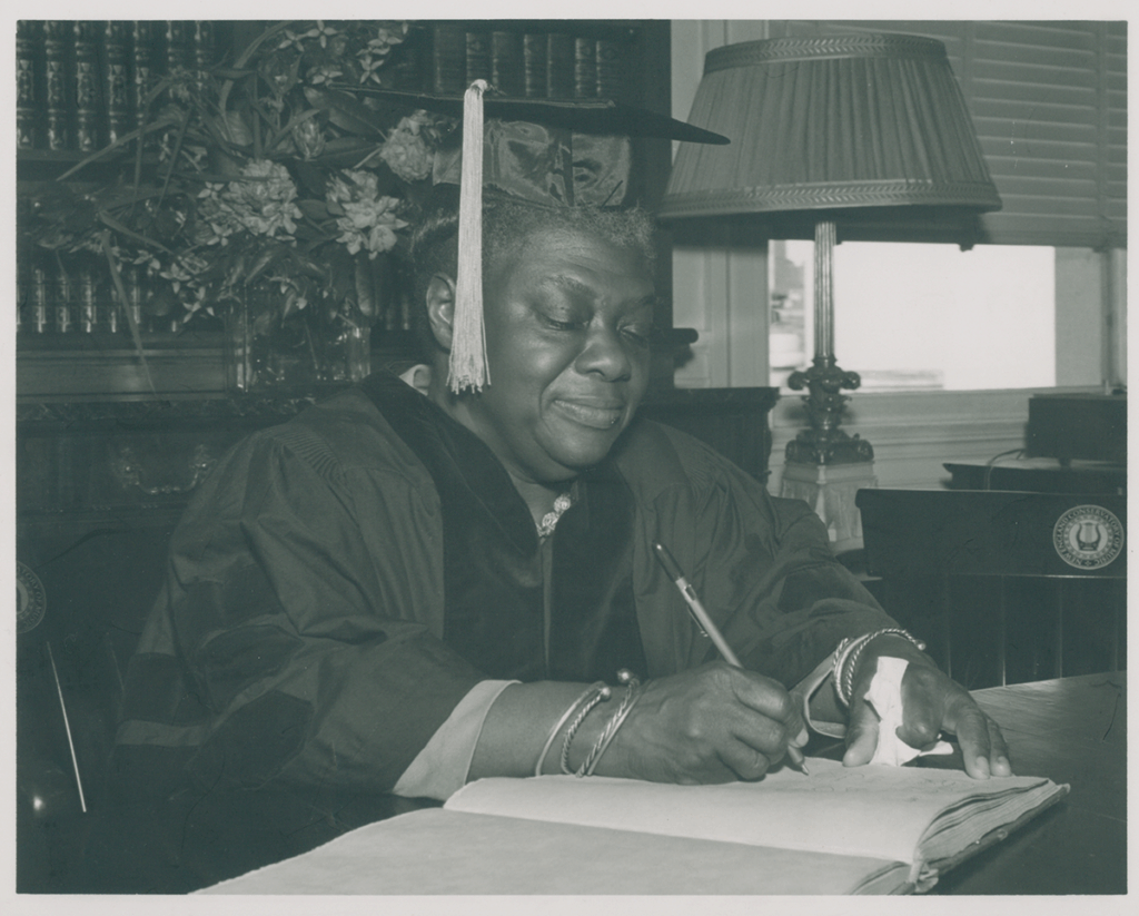 Black and white image of Elma Lewis writing at a desk while wearing a cap and gown