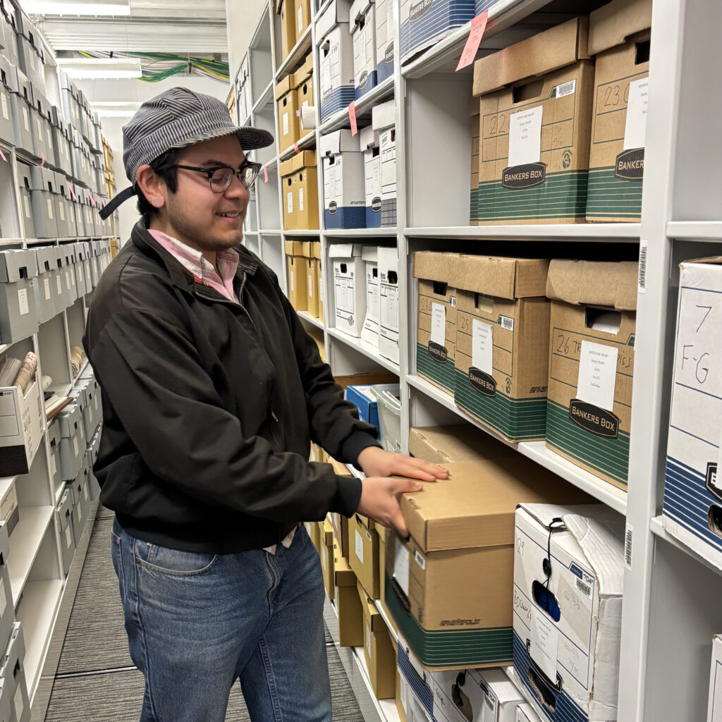 Aries Peralta, wearing a black jacket, gray baseball cap, and glasses, pulls a box off a shelf in the archives