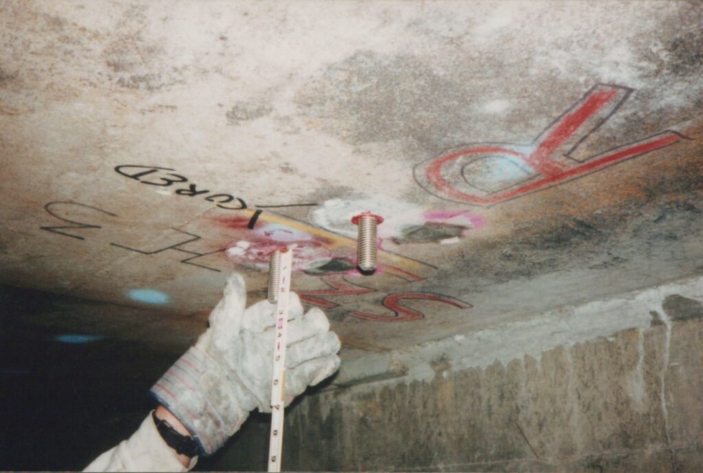 A gloved hand holds a tape measure to a concrete ceiling, measuring the length of screws sticking down.