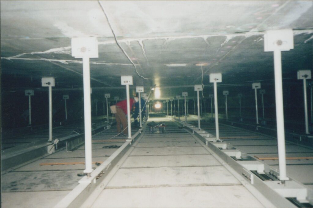 A worker stands in a crawlspace above the ceiling of a tunnel, surrounded by concrete and bars.