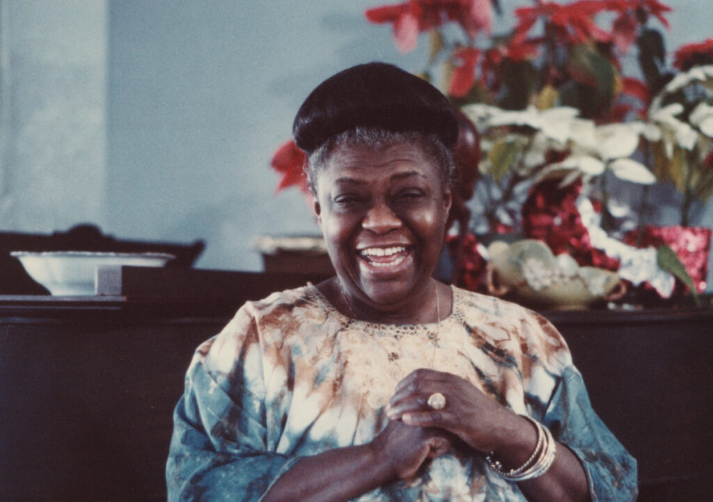 Elma Lewis, a Black woman with dark hair in a bun, stands in front of flowers with a huge smile on her face. Her hands are clasped over her chest.