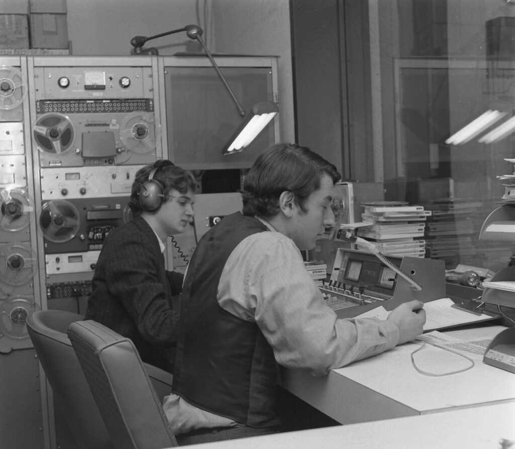 Black and white image of two students sitting in a recording studio. They are wearing headphones and sitting at a table while surrounded by 1960s-era recording equipment