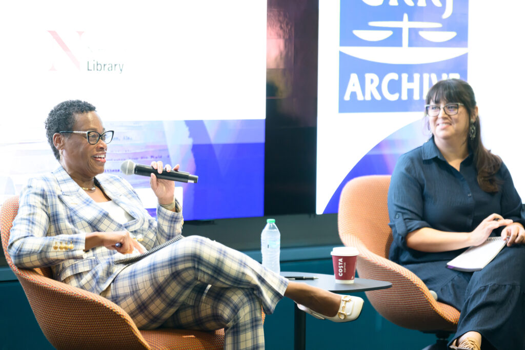 Two women sit in front of a large screen. One is holding a microphone and speaking