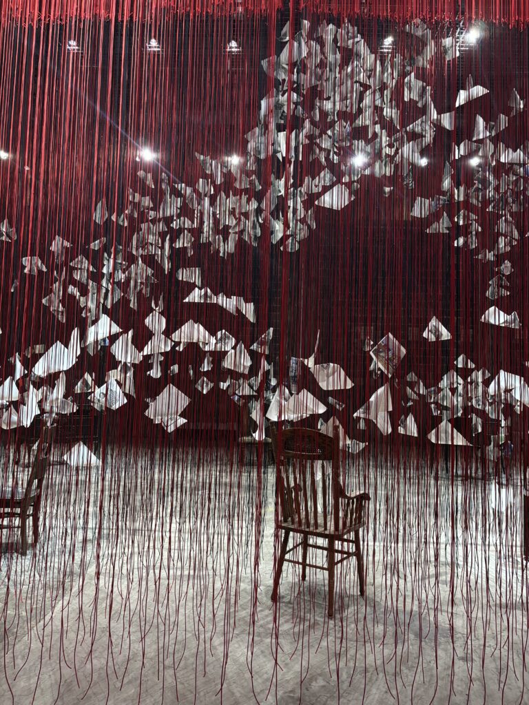 A series of red and black threads hanging from the ceiling with folded papers suspended within. Two chairs also sit within the threads