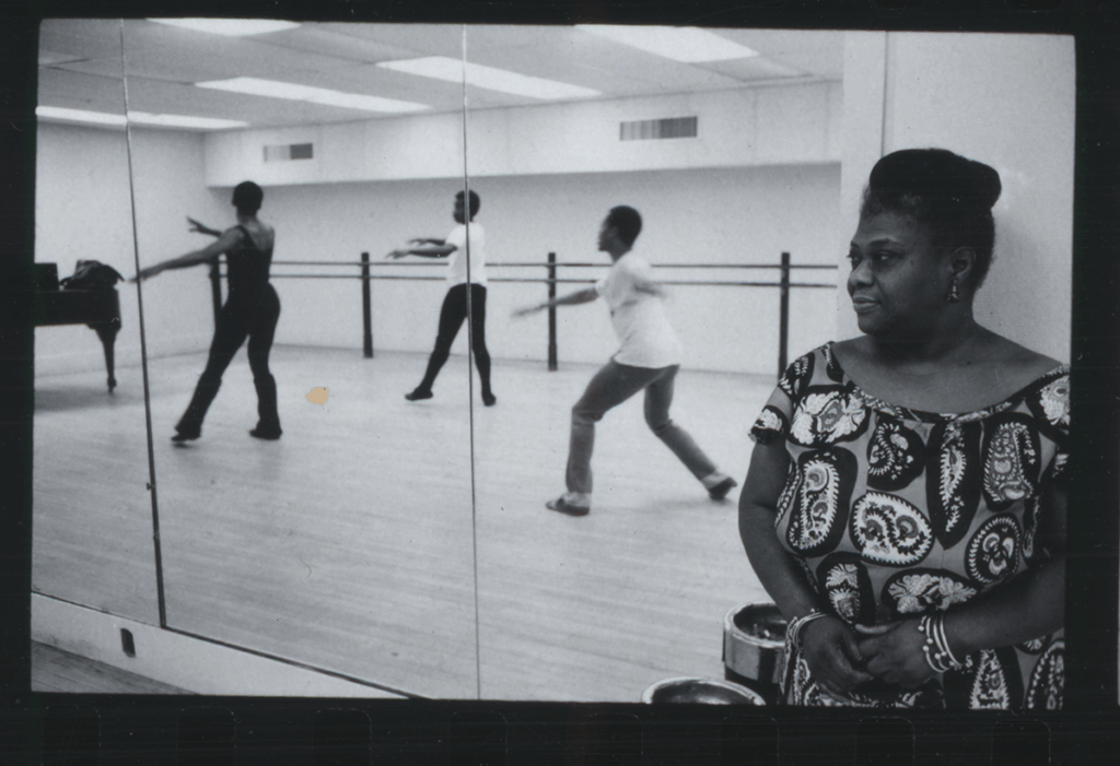Black and white image of a woman wearing a dress and with her hair in a bun watching three dancers in a dance room. The dancers are visible behind her in a mirror