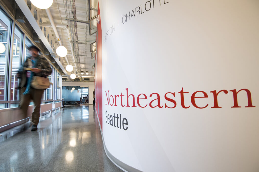 A blurry person walks by a curved white wall with "Northeastern Seattle" written on it.