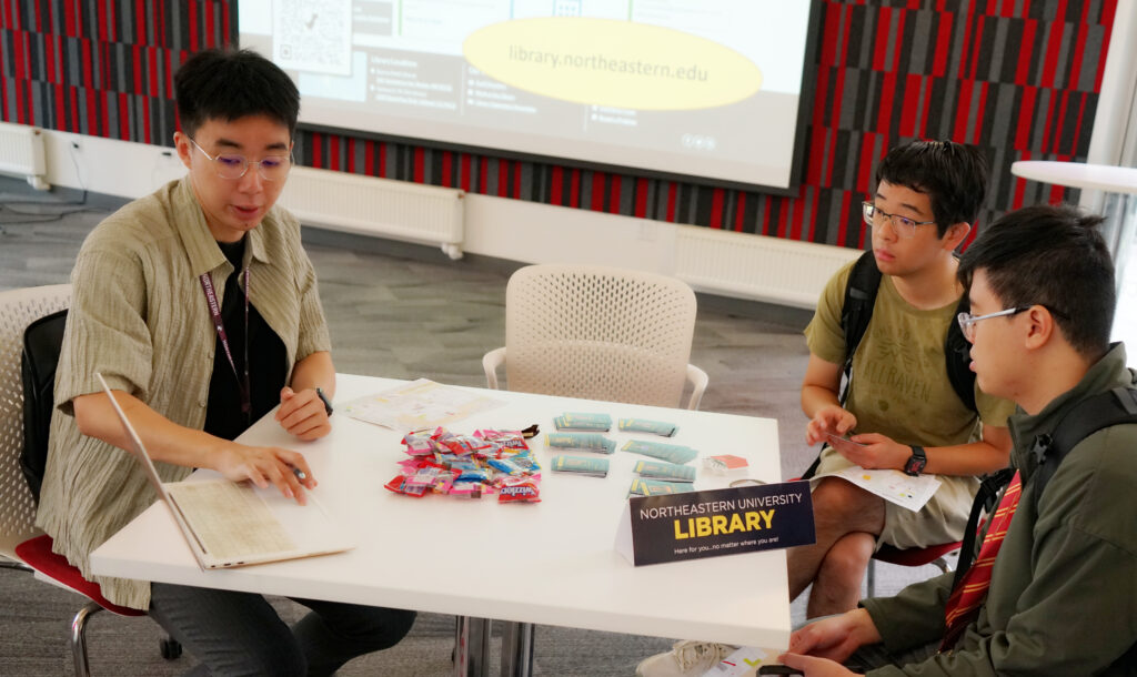 Three students sit around a small table. The one on the left (Jason Wu) is pointing to a laptop. The laptop is facing the other side of the table, where two students listen.