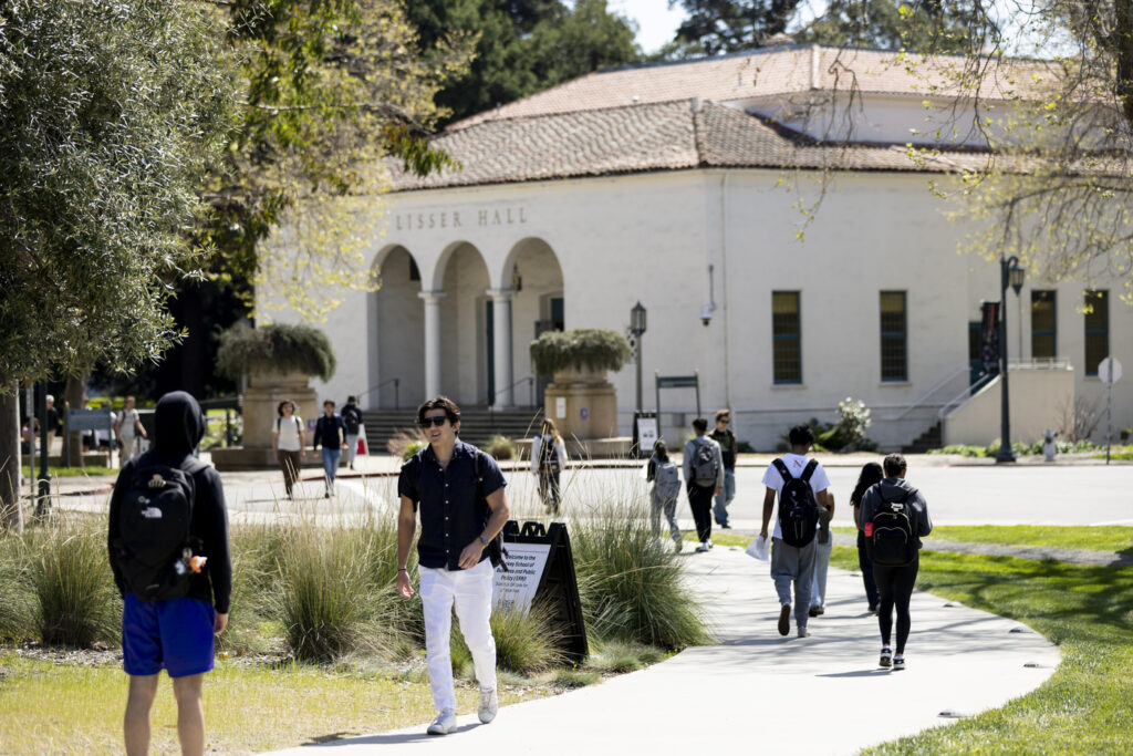 Several students walk around campus paths on a sunny day. A white building with a sign that says "LISSER HALL" is in the background