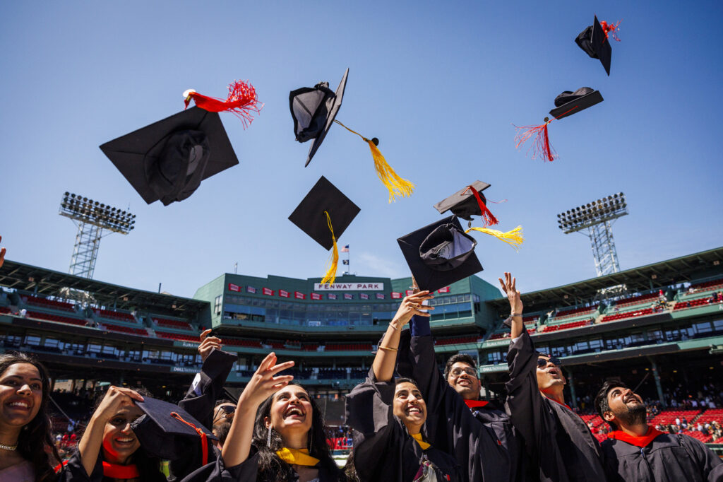 Graduates throw their caps in the air under a blue sky in Fenway Park. The Fenway Park sign is visible behind the caps
