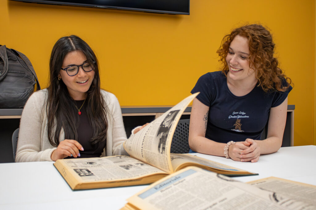 Two students sit at a table flipping through a bound collection of newspapers
