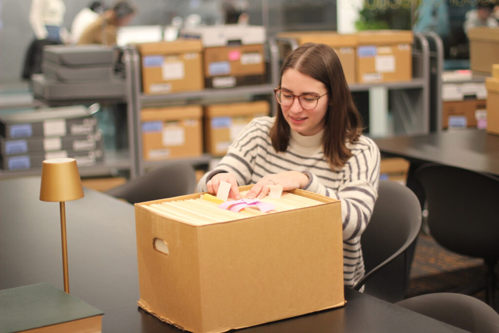 A person sitting at a table in the Snell Library Archives Reading Room flips through files in an archival box. More boxes are in the background.