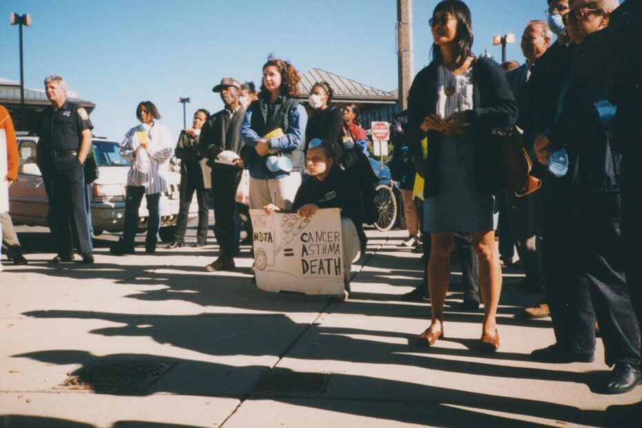 A group of protestors stand on a sidewalk, one holding a sign that reads "CANCER = ASTHMA DEATH"